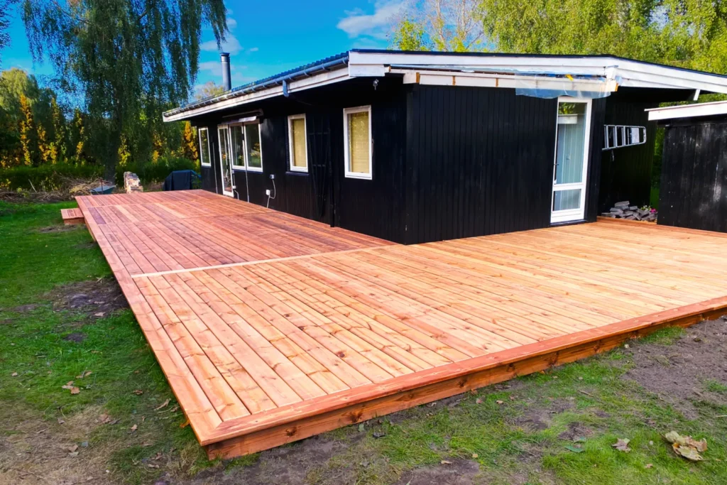 Den store træterrasse i vinkel omkring huset, hvor terrassen samler udgangen fra stuen og den vestvendte facade på sommerhuset. Trappen åbner mod haven i Rågeleje.